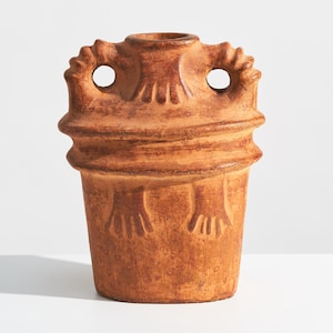 Puede incluir: Una vasija de cerámica color terracota con base cilíndrica y una banda horizontal ancha y elevada en el centro. La parte superior presenta dos asas laterales con detalles estriados y una abertura central, con motivos en forma de manos en el cuerpo.