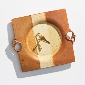 Op de afbeelding: Vierkante keramische schaal met een centrale cirkelvormige inkeping. De schaal heeft een colorblock-ontwerp met een bruin en crèmekleurig kleurenschema. Twee sleutels liggen in de cirkelvormige inkeping. De schaal heeft twee decoratieve handvatten.