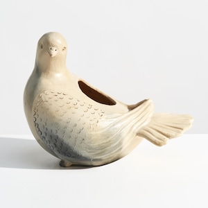 May include: A ceramic bird-shaped planter with a matte beige finish and subtle grey shading on the wings and chest. The sculpture features carved feather details, a smooth head, and an open cavity on the back for holding small items or plants.