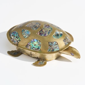 Op de afbeelding: Een messing schildpadbeeldje met een schild ingelegd met iriserende abalone stukken. De schildpad heeft een gedetailleerd ontwerp, met zichtbare poten, kop en staart. Het beeldje is een decoratief item.