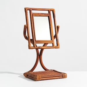 May include: A rectangular tabletop mirror framed in brown rattan. The mirror is suspended within a matching rattan frame that pivots on two curved supports, which are attached to a square base featuring a woven rattan surface texture.