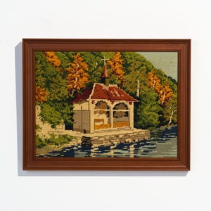 May include: Framed needlepoint art depicting a lakeside building with a red roof, surrounded by trees with green and orange foliage. The artwork is in a brown wooden frame, creating a classic and detailed landscape scene.