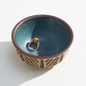 May include: A small, round ceramic bowl with a dark brown exterior featuring vertical carved grooves and a smooth, matte teal interior. Resting inside the bowl is a gold ring with a rectangular gemstone setting, catching the light.