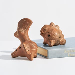 May include: Two brown ceramic figurines. One is a stylized bird-like figure with a long neck and a rounded body. The other is a small, rounded animal with a textured surface. Both are placed on a light blue book.