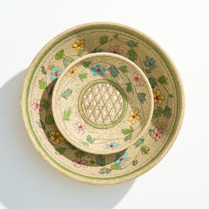 Könnte beinhalten: Zwei beige Keramikschalen mit floralen Designs. Die größere Schale hat einen grünen Rand und ist mit bunten Blumen und grünen Blättern verziert. Die kleinere Schale, die sich darin befindet, hat ein ähnliches Design mit einem Rautenmuster in der Mitte.