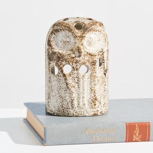 May include: A cylindrical ceramic owl figurine with a speckled brown and cream glaze. The owl features cut-out circular eye and wing details, with a flat top containing small circular openings. It rests on a light blue book titled American Drama.