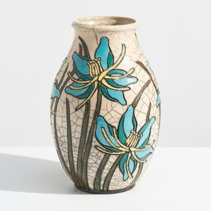May include: A ceramic vase with a cream-colored crackle glaze finish, featuring hand-painted blue flowers with yellow centers and green stems. The vase has an oval body, a narrow neck, and a flared rim, showcasing a detailed floral motif.