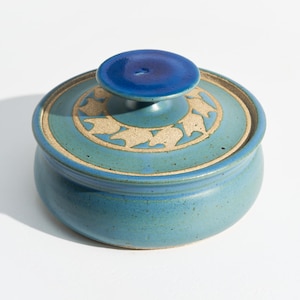 May include: A round ceramic lidded container with a speckled teal blue glaze. The lid features a central dark blue knob and a circular band of light beige leaf-shaped patterns embossed into the surface, contrasting with the smooth blue finish.