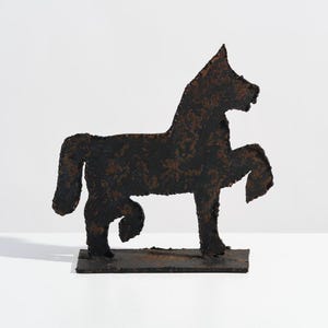 Puede incluir: Una escultura de metal negro de un caballo de pie sobre una pequeña base. La escultura está hecha de metal rugoso y texturizado y tiene un aspecto rústico.