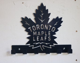 May include: Black metal key holder shaped like a Toronto Maple Leafs logo. The logo is a stylized maple leaf with the team's name written inside. The key holder has six hooks.