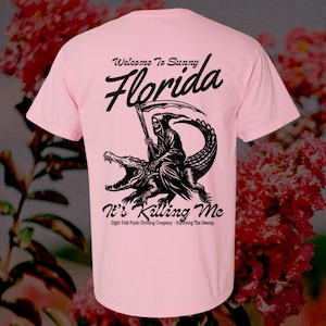 Peut inclure: T-shirt rose avec un graphique noir d'un Grim Reaper chevauchant un alligator. Le texte "Welcome to Sunny Florida" et "It's Killing Me" est imprimé sur le t-shirt.