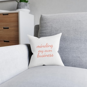 Peut inclure: Un coussin carré blanc avec l'inscription "minding my own business" en script corail. Le coussin est posé sur un canapé gris, avec un fond neutre.
