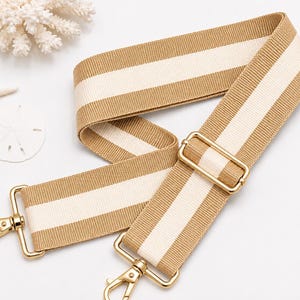 May include: A woven bag strap featuring a wide beige and cream striped pattern. The strap includes gold-toned metal hardware, including two swivel lobster clasps at the ends and an adjustable sliding buckle to modify the length of the strap.