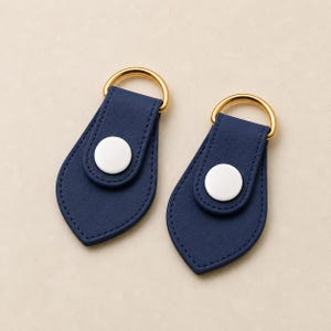 May include: Two navy blue teardrop-shaped leather tabs with gold-toned metal D-rings at the top. Each tab features a centered white circular snap button closure and matching navy blue stitching along the edges of the folded leather material.