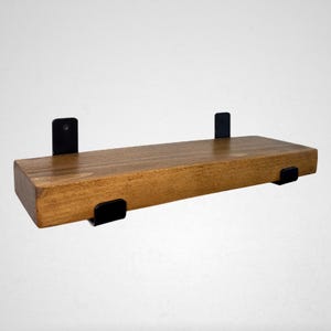 May include: A rectangular wooden shelf with a warm brown finish. The shelf is supported by four black metal brackets, two on the top and two on the bottom. The shelf is designed for wall mounting and is suitable for displaying items.