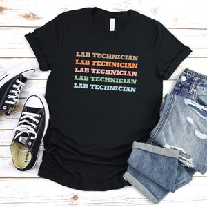 Puede incluir: Camiseta negra con el texto "Lab Technician" repetido cinco veces en colores arcoíris. La camiseta está colocada sobre una superficie de madera blanca con un par de zapatillas negras y unos vaqueros azules.
