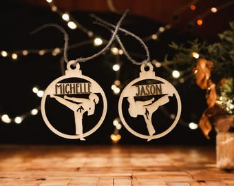 May include: Two wooden Christmas ornaments with a karate theme. Each ornament is circular with a cutout of a karate figure and a name: "MICHELLE" and "JASON". The ornaments are hanging against a background of fairy lights.
