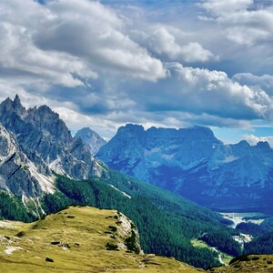 May include: A panoramic view of a mountain range with lush green valleys and a blue sky with white clouds. The mountains are rocky and jagged, with some snow-capped peaks. The valley is covered in trees and there is a river winding through it.