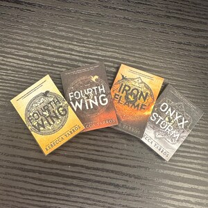 May include: Four fantasy novel book covers with black and gold lettering on a dark wood background. The titles are "Fourth Wing", "Iron Flame", "Onyx Storm", and "Fourth Wing".
