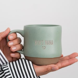 Puede incluir: Una taza de cerámica verde salvia con acabado moteado y base marrón lisa. El frente de la taza presenta el texto grabado "BOB'S NANA" en una fuente sencilla, situado sobre un pequeño contorno de corazón grabado y centrado en el cuerpo.