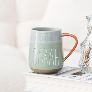 May include: A ceramic mug with the name "SARAH" in white lettering. The mug has a light green body, a gray rim, and a brown handle. It sits on a stack of books next to a glass vase. A business card holder with the text "Oneup custom ceramic gifts" is in the foreground.