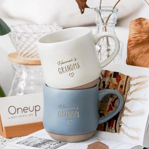 May include: Two ceramic mugs stacked vertically. The top mug is off-white with speckles and features the text Hannah's GRANDMA with a heart. The bottom mug is blue with speckles and features the text Hannah's GRANDPA with a heart. Both have handles.
