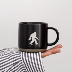 May include: Black coffee mug with a Bigfoot silhouette. The mug has a dark exterior with a contrasting brown base and a black handle. The mug is held by a person wearing a striped shirt.