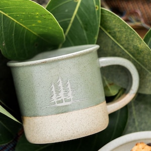 Puede incluir: Una taza de cerámica verde salvia con base y asa color crema. La taza presenta un diseño de árbol blanco y está rodeada de hojas verdes. Un plato de galletas con chispas de chocolate está en primer plano.
