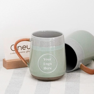 May include: Two ceramic mugs with a light green and gray color scheme. The mugs have a brown handle and a white circle with the text "Your Logo Here". One mug is upright, and the other is on its side. The mugs are on a striped surface.