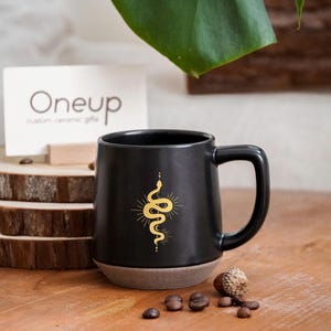 Peut inclure: Mug en céramique noire avec un motif de serpent et de soleil doré. Le mug a une base marron et une grande anse. Le mug est posé sur une surface en bois avec des grains de café et un gland.