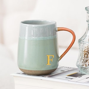 Puede incluir: Una taza de cerámica con cuerpo verde claro, borde gris y asa color cobre. La letra "F" es dorada en la taza. La taza está sobre una pila de libros junto a un jarrón de cristal. Un tarjetero con el texto "Oneup custom ceramic gifts" está en primer plano.