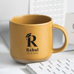 May include: Mustard yellow ceramic mug with a gray base. The mug features a black graduation cap and the letter "R", with the name "Rahul" and "Ph.D. 2025" printed below. A white keyboard and mouse are in the foreground.