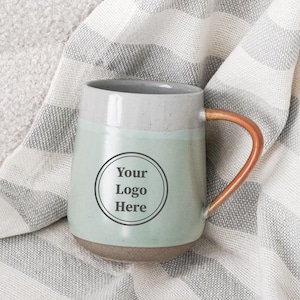 May include: A ceramic mug with a light green base and a gray top, featuring a copper-colored handle. The mug has a circular logo area with the text "Your Logo Here". It rests on a striped gray and white blanket.