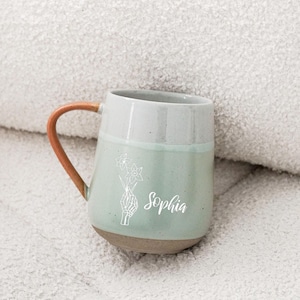 Puede incluir: Una taza de cerámica con un esmalte verde claro y gris, con un asa marrón. La taza tiene el nombre "Sophia" y un diseño floral impreso en la parte delantera. La taza está sobre una superficie blanca texturizada.