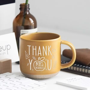Könnte beinhalten: Senfgelbe Keramiktasse mit den weißen Worten "THANK YOU MRS WARREN". Ein grünes Monstera-Blatt befindet sich in einer braunen Glasflasche. Eine weiße Tastatur und ein Kartenhalter aus Holz befinden sich ebenfalls auf der weißen Oberfläche.