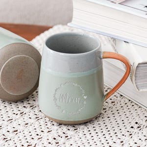Puede incluir: Dos tazas de cerámica en tonos verde claro y gris, con asa marrón. Una taza muestra la palabra "Mom" dentro de una corona floral. La otra taza está tumbada. Un letrero dice "Oneup custom ceramic gifts".