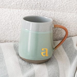 Puede incluir: Una taza de cerámica verde menta y gris con asa y base marrón. La taza tiene la letra dorada "a" en la parte delantera. La taza está sobre una tela a rayas, sobre un fondo blanco texturizado.