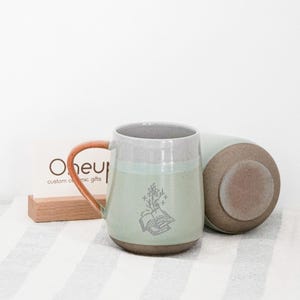 Puede incluir: Dos tazas de cerámica, una en posición vertical, la otra de lado. La taza vertical es de color verde claro con la parte superior gris y un asa marrón. Un diseño floral y de libro está en la parte delantera. La otra taza es verde claro y marrón. Una tarjeta de visita está en el fondo.