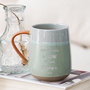 Puede incluir: Una taza de cerámica con un esmalte verde claro y gris, con un asa marrón y la inscripción "May your coffee be STRONG and your Monday be short." La taza está sobre una pila de libros, con una botella de vidrio transparente en el fondo.
