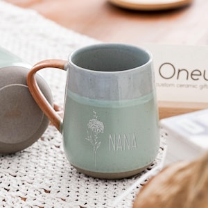 May include: A ceramic mug with a light green body and a brown handle. The mug has the word "NANA" and a floral design etched on the front. The mug is part of a set of custom ceramic gifts.