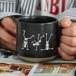 Könnte beinhalten: Schwarze Keramik-Tasse mit beigefarbenem Boden, verziert mit weißen Skelett-Illustrationen. Die Tasse wird über einem Magazin mit farbigen Bildern gehalten. Die Person, die die Tasse hält, trägt ein gestreiftes Hemd.