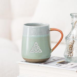 Puede incluir: Una taza de cerámica con cuerpo verde claro, borde gris y asa marrón. La taza presenta un diseño de nudo celta blanco. La taza está sobre una pila de libros junto a un pequeño jarrón de cristal. Un tarjetero con el texto "Oneup custom ceramic gifts" está en primer plano.