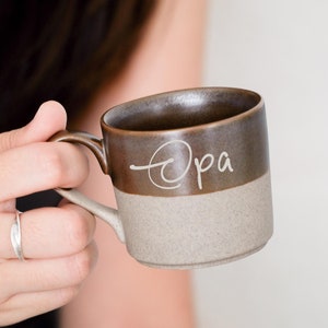 May include: A brown and beige ceramic mug with the word "opa" written on it in cursive.