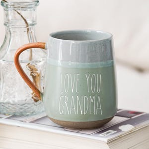 Puede incluir: Una taza de cerámica de dos tonos con el texto blanco "LOVE YOU GRANDMA". La taza tiene un cuerpo verde claro y una parte superior gris, con un asa marrón. Descansa sobre una pila de libros. A la izquierda hay una botella de vidrio transparente.