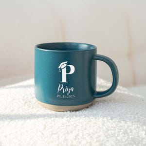 May include: Teal ceramic mug with a white graduation cap and the letter "P" design. The name "Priya" and "Ph.D. 2025" are printed below the letter. The mug has a beige base and a curved handle.