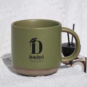 May include: A green ceramic mug with a speckled finish and a tan base. The mug features a black graduation cap graphic with the letter "D" and the name "Dmitri" and "Ph.D. 2025" printed below. The mug has a curved handle.