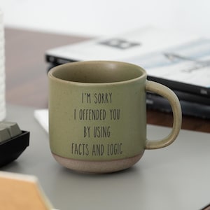 Pode incluir: Uma caneca de cerâmica verde-oliva com o texto "I'M SORRY I OFFENDED YOU BY USING FACTS AND LOGIC". A caneca tem uma base bege e uma alça curva. Um teclado e material de escritório estão no fundo.