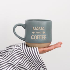 Puede incluir: Una taza de café de cerámica color turquesa con base beige, sostenida en una mano. La taza tiene un asa curva y las palabras "MAMA NEEDS COFFEE" impresas en la parte delantera en color beige claro. La persona lleva una camisa a rayas blancas y negras.