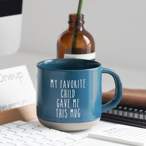 Puede incluir: Una taza de cerámica color turquesa con el texto "MY FAVORITE CHILD GAVE ME THIS MUG" en blanco. La taza tiene una base gris y un asa curvada. Una planta verde y una pequeña botella marrón están en el fondo.