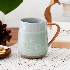 May include: A ceramic mug with a light green body and a gray rim. The mug features the word "MOM" and a floral design. It has a brown handle and sits on a white lace surface. Other items in the background include a wooden plate with cookies.